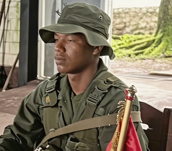 Hombre con uniforme militar verde, sombrero táctico y arnés de combate junto a un asta dorada con tela roja