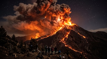 Un volcán en erupción expulsa lava y ceniza durante la noche mientras personas en silueta observan la escena desde una colina.