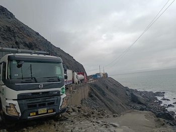 Camiones y vehículos de carga pesada varados en una carretera costera. A un lado, una ladera rocosa; al otro, el mar. Cielo nublado