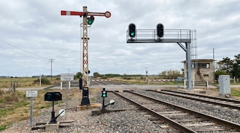 Imagen de un paso a nivel con vías de tren, varias señales ferroviarias, semáforos en verde, una barrera de seguridad y un paisaje rural bajo cielo nublado.