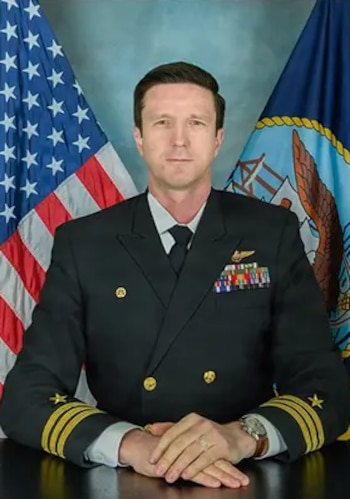 Retrato de un hombre de mediana edad vestido con uniforme militar azul marino, insignias y medallas. Banderas de Estados Unidos y de la Armada de fondo