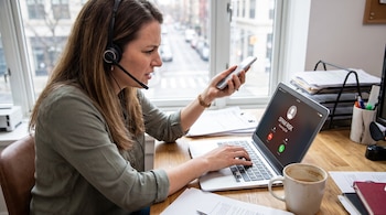 Mujer con auriculares y micrófono sostiene un teléfono móvil, mientras mira un ordenador portátil que muestra una llamada de riesgo spam en un escritorio.