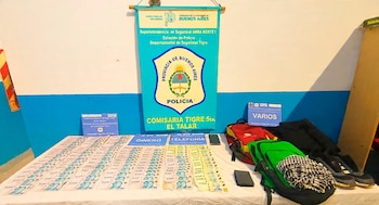 Mesa con billetes, teléfonos, mochilas y prendas de vestir frente a un cartel de la Comisaría Tigre 5ta. El Talar de la Policía de Buenos Aires