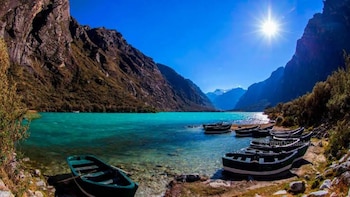 Lago de agua turquesa con varias barcas de madera a la orilla y dentro del agua, rodeado de montañas imponentes bajo un cielo azul soleado