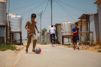 Dos niños juegan fútbol en una calle de un campamento. Uno dribla un balón rojo y azul; el otro corre cerca. Tanques de agua y edificios simples se ven al fondo