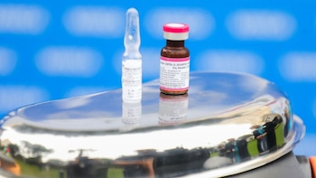 Dos viales de medicamento, uno transparente y otro marrón con tapa rosa, se reflejan en una superficie metálica. El vial marrón es una vacuna contra el sarampión sobre fondo azul