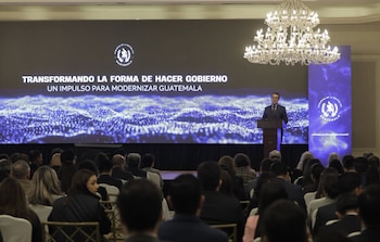 Hombre en podio habla frente a pantalla grande con texto "Transformando la forma de hacer gobierno" y público sentado en auditorio. Candelabro superior