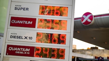 Tablero digital blanco con precios de combustible Axion SUPER, QUANTUM y DIESEL X10 en números rojos. Fondo borroso con un logo circular rojo y blanco