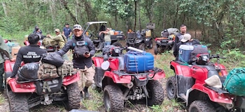 Varias personas permanecen junto a cuatrimotos rojas y equipo de expedición en un área boscosa rodeada de vegetación