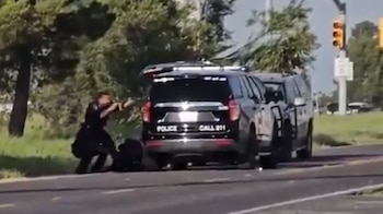 Un oficial de policía con uniforme oscuro se agacha detrás de una camioneta SUV negra con insignias 'POLICE' y 'CALL 911' en la parte trasera, apuntando un arma hacia adelante