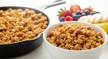 Granola dorada y crocante de avena y frutos secos en un sartén de hierro fundido, y servida en un bowl blanco. Al fondo, un plato con fresas, arándanos y plátano.