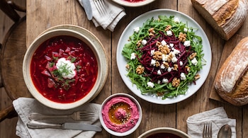 Mesa de madera con sopa borscht, ensalada de rúcula, remolacha rallada, nueces, trozos de queso blanco, hummus de remolacha, pan, cubiertos y servilletas.