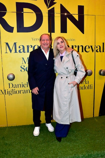 Un hombre con abrigo oscuro y una mujer con gabardina beige posan sonrientes frente a un panel amarillo con letras y césped artificial
