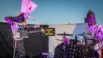 Dos músicos con trajes y máscaras de lunares blancos y negros, uno toca guitarra eléctrica y el otro batería. Detrás, un letrero dice "Angine de Poitrine"