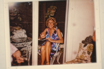 Fotografía de Yiya Murano, una mujer rubia sonriente sentada en una silla de jardín blanca con un vestido de colores, levantando la mano derecha