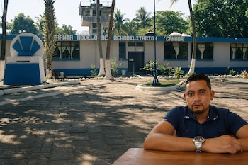 Un hombre con camisa azul sentado frente a una mesa con el patio y la fachada de un centro de rehabilitación al fondo.
