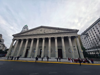 Vista frontal de la Catedral Metropolitana de Buenos Aires con sus columnas, friso y personas en la entrada, bajo un cielo nublado, con banderas celestes y blancas