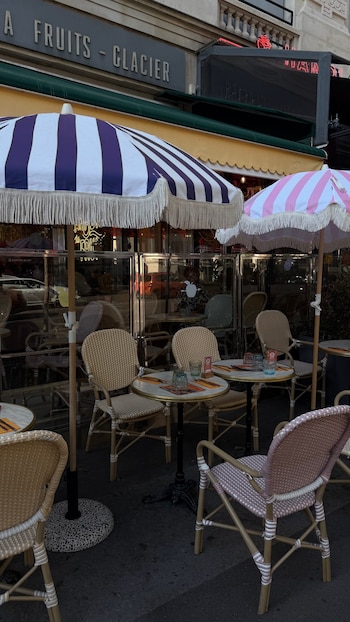 Terraza exterior con mesas redondas de mármol, sillas de mimbre, dos sombrillas a rayas blancas y púrpuras, y blancas y rosas, frente a una fachada de local con cartel