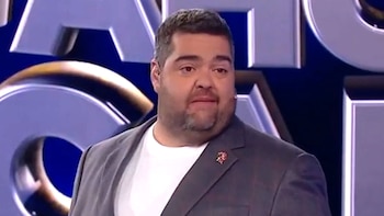 Darío Barassi, un hombre con barba, camiseta blanca y saco gris, muestra una expresión de perplejidad frente a un fondo oscuro con detalles metálicos