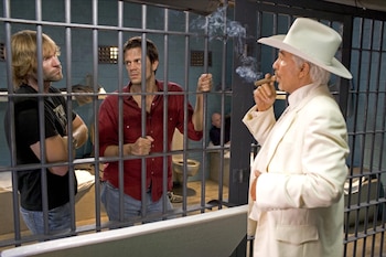 Burt Reynolds fuma un cigarro, observando a Johnny Knoxville y Seann William Scott tras los barrotes de una celda de prisión con un hombre al fondo