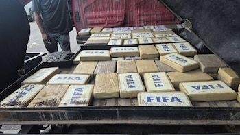 Parte trasera de un camión rojo cargada con numerosos paquetes rectangulares de color beige, algunos marcados con "FIFA", y un neumático