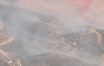 Vista aérea de colinas áridas quemadas con humo blanco elevándose y algunas zonas rojas, con líneas eléctricas y un camino de tierra sinuoso