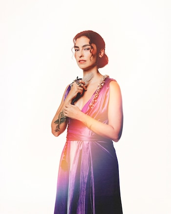 Una mujer posa frente a un fondo blanco con un vestido largo de color morado brillante, el cabello recogido y un tatuaje en el antebrazo