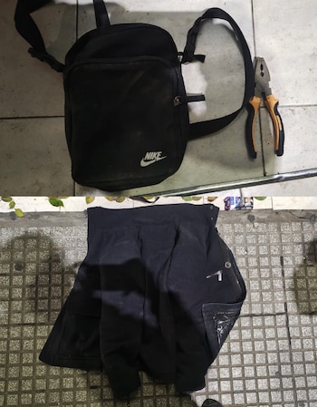 Una bolsa de hombro negra con logo Nike, unos alicates de mango naranja y una chaqueta negra doblada sobre un suelo de baldosas grises