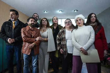 Un grupo de ocho personas, incluyendo hombres y mujeres de diversas edades, de pie en un interior. Visten ropa casual y formal, mirando hacia arriba o al frente
