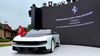 Un hombre de traje y sombrero rojo posa junto a un coche blanco en un evento al aire libre. Detrás, una pantalla negra muestra el logo de Ferrari y texto sobre el vehículo