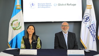 Dos personas, una mujer y un hombre, sentadas en una mesa frente a banderas de Guatemala y una pantalla con logos de Bienestar Social y Global Refuge