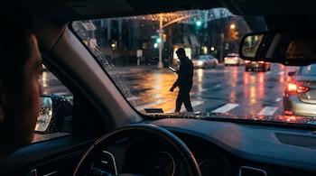 Vista desde el interior de un vehículo hacia un hombre que cruza la calle bajo la lluvia mientras mira la pantalla de un teléfono.