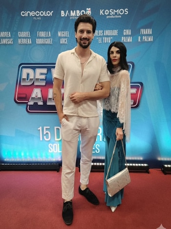 Fiorella Rodríguez e Ivan Micol posan de pie en una alfombra roja. Él viste camisa y pantalón claro, ella blusa oscura, capa de encaje blanca y pantalón azul