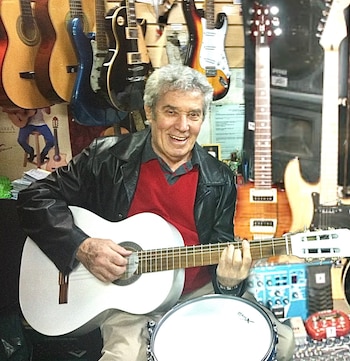 Un hombre de cabello gris sonríe y toca una guitarra acústica blanca. Detrás hay guitarras eléctricas y acústicas colgando en una tienda de instrumentos