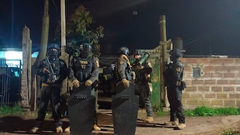 Cuatro agentes de fuerzas especiales con equipo táctico, cascos, máscaras y armas, posan con escudos y una herramienta de entrada, de noche en un terreno irregular