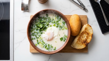 Plano cenital de changua colombiana humeante en tazón de madera con huevo, cilantro y cebolla, junto a almojábana y calado en tabla de madera.