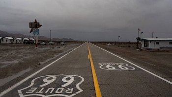 Carretera desértica, la Ruta 66, con charcos de agua y el número "66" pintado en el asfalto, bajo un cielo nublado, con el Roy's Motel y luces de la calle
