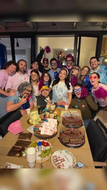 Grupo de personas posando alrededor de una mesa de madera con comida, dulces, bebidas, un pastel con globos, platos y decoraciones de fiesta