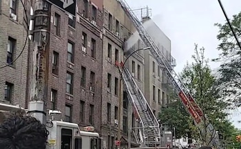 Una escalera de bomberos se extiende hacia un edificio de ladrillo de varios pisos con humo saliendo de una ventana superior, mientras un camión de bomberos está en la calle