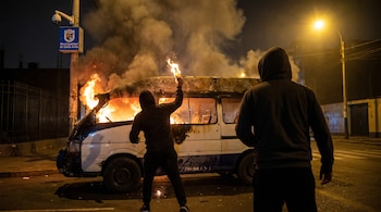Una combi blanca y azul en llamas por dos figuras encapuchadas en la noche. Un atacante lanza un objeto incendiario, el otro observa, con una cámara de seguridad de Santa Anita al fondo.