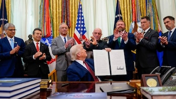 Un hombre de pelo rubio sentado en un escritorio sostiene un documento abierto, mientras varios hombres lo rodean y aplauden. Hay banderas estadounidenses y cintas de colores detrás