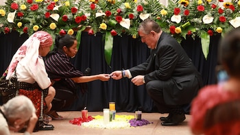 Um homem de terno e duas mulheres com roupas tradicionais curvando-se diante de um altar com velas, flores e líquido em copos, com fundo floral