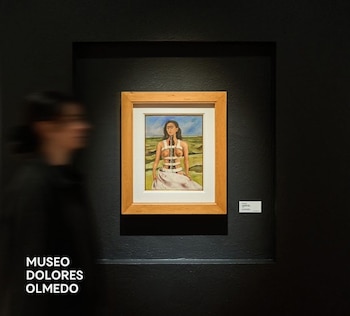 Pintura enmarcada de Frida Kahlo en una pared oscura de museo, con una etiqueta y una persona borrosa caminando. Texto: MUSEO DOLORES OLMEDO