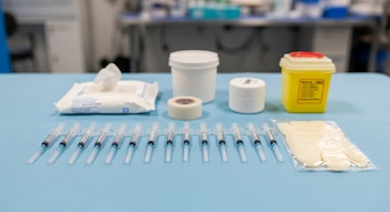 En una mesa azul, diez jeringas prellenadas se alinean junto a toallitas, cinta médica, dos recipientes blancos, un contenedor amarillo de objetos punzantes y guantes.