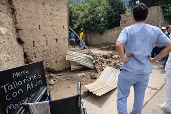 Vista trasera de persona en uniforme médico azul mirando un área dañada con paredes de adobe y escombros. En primer plano, cartel de menú y silla de ruedas rota.