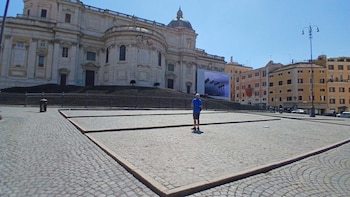 Gran basílica blanca con escaleras en Roma bajo cielo azul, un hombre de espaldas en el centro de una plaza adoquinada