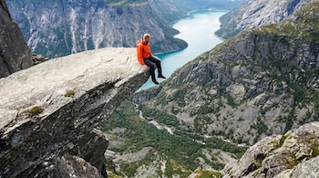 Un hombre calvo con chaqueta naranja y pantalón oscuro está sentado en el borde de una roca que sobresale, con un gran fiordo turquesa y montañas al fondo.