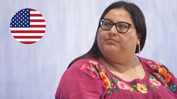 Mujer con cabello oscuro, gafas, blusa con bordados florales de colores y aretes dorados; bandera de Estados Unidos en círculo.