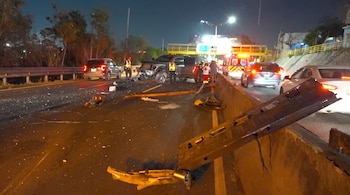 Carretera nocturna con dos vehículos colisionados, uno tipo camioneta y otro automóvil, rodeados de personal de emergencia y un vehículo de ambulancia con luces encendidas. Escombros dispersos.