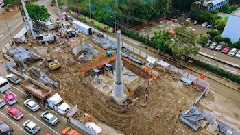 Vista aérea de una obra con una columna central de concreto, maquinaria pesada, excavaciones y una avenida con vehículos al costado